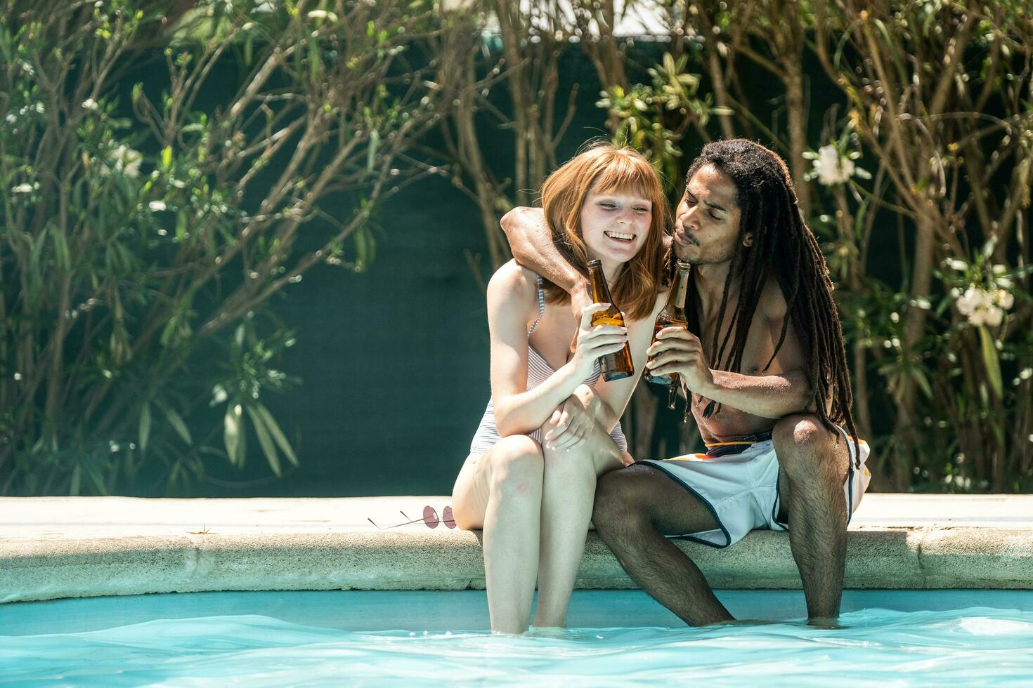 afro-americano homem e branco mulher brindar com Cerveja garrafas em a Beira do uma piscina. foto