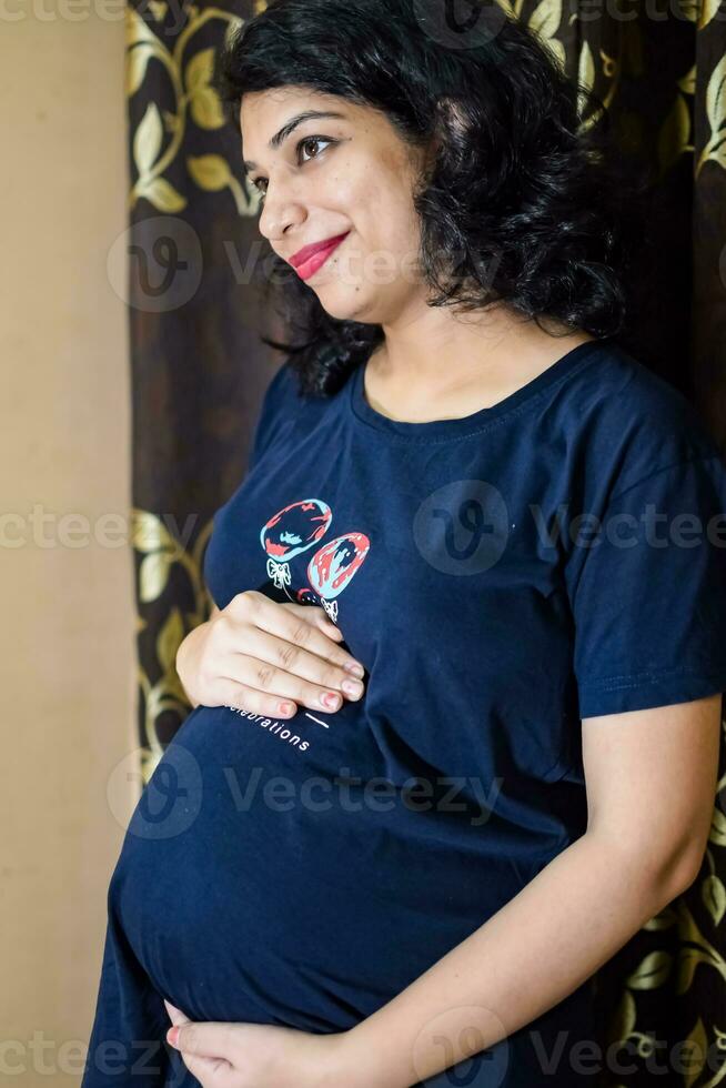 uma grávida indiano senhora poses para interior gravidez tiro e mãos em barriga, indiano grávida mulher coloca dela mão em dela estômago com uma maternidade vestir dentro dela casa foto