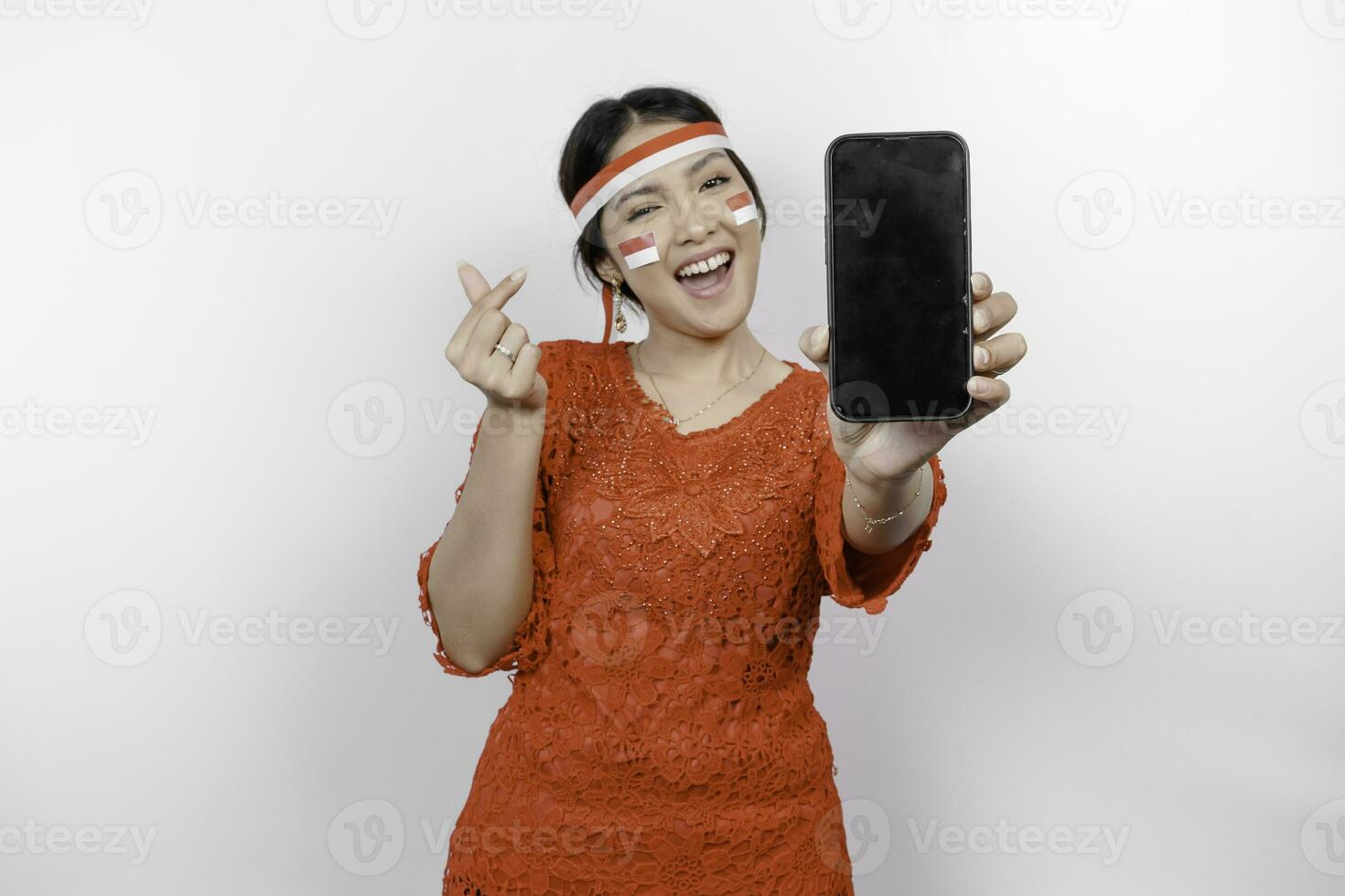 uma feliz ásia mulher vestindo vermelho kebaya e arco de cabelo, mostrando dela telefone, e gesticulando amor placa de dela dedos mostrando concurso sentimento, isolado de branco fundo. da indonésia independência dia foto