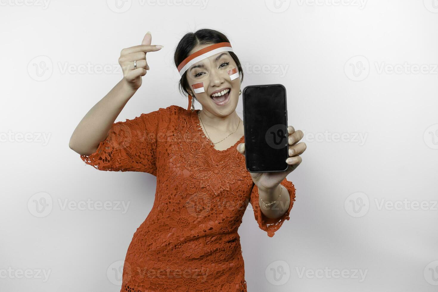 uma feliz ásia mulher vestindo vermelho kebaya e arco de cabelo, mostrando dela telefone, e gesticulando amor placa de dela dedos mostrando concurso sentimento, isolado de branco fundo. da indonésia independência dia foto