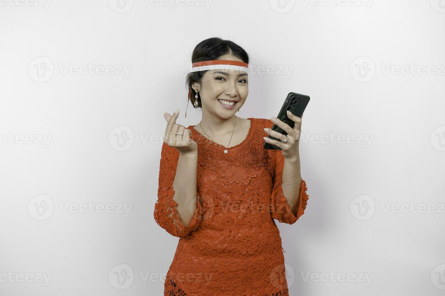 uma feliz ásia mulher vestindo vermelho kebaya e arco de cabelo, segurando dela telefone, e gesticulando amor placa de dela dedos mostrando concurso sentimento, isolado de branco fundo. da indonésia independência dia foto