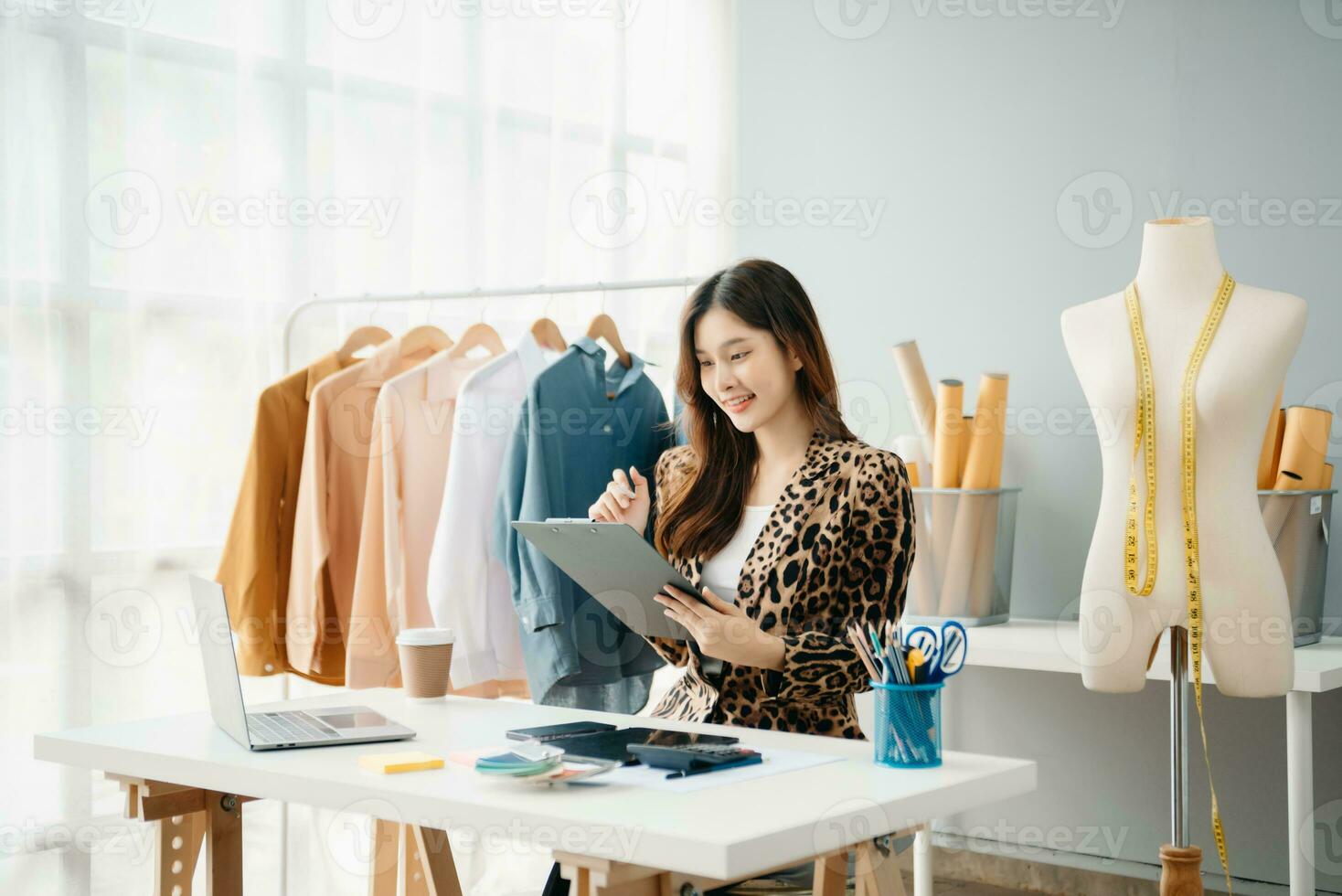 ásia jovem mulher em escrivaninha dentro escritório do moda desenhador e detém tábua, computador portátil e Smartphone foto