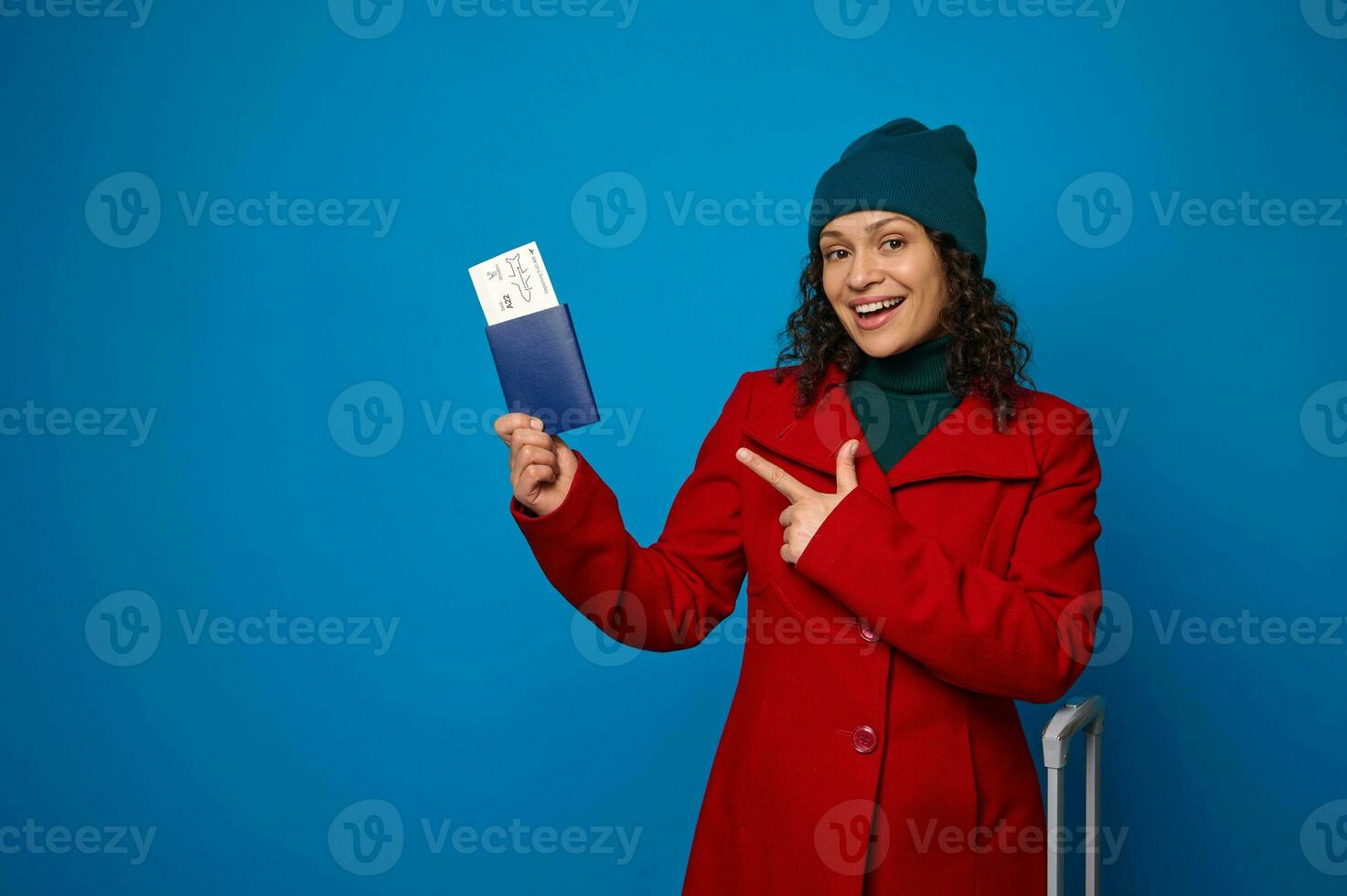 jovem sorridente africano mulher dentro brilhante vermelho casaco, verde caloroso de lã chapéu, pontos em Passaporte com ar bilhete e embarque passar dentro mãos. viagem, ar voar viagem conceito em azul fundo, cópia de espaço foto