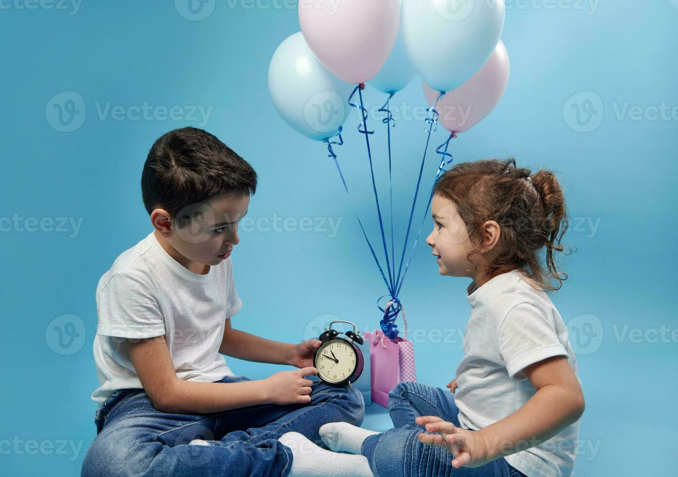 pequeno Garoto segurando a alarme relógio e mostrando isto para dele irmã em a fundo do colori balões. foto