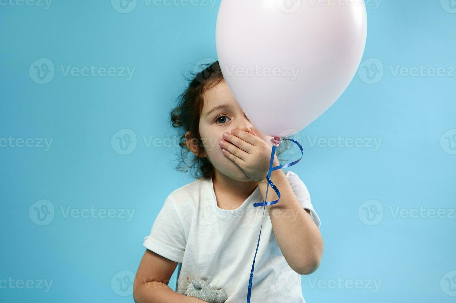 adorável criança menina cobre dela boca com dela mão e poses para Câmera atrás uma Rosa balão em azul fundo com cópia de espaço foto