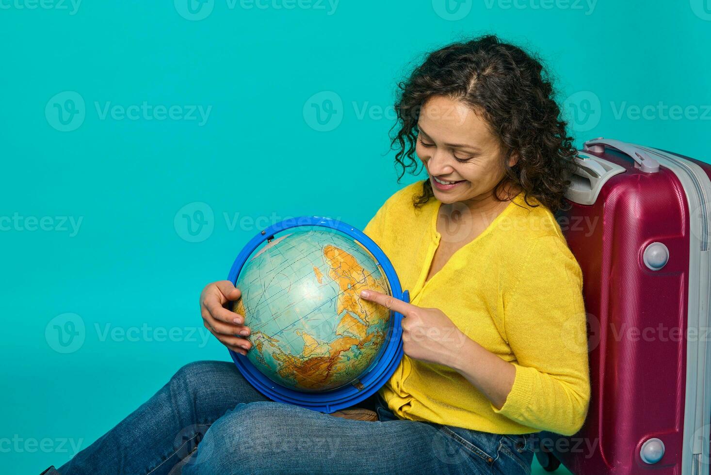 encantador mulher viajante dentro amarelo camisa senta Próximo para uma mala de viagem e pontos com dela dedo em uma Lugar, colocar em a globo, escolhendo uma viagem destino. turismo conceito em azul fundo com cópia de de Anúncios espaço foto