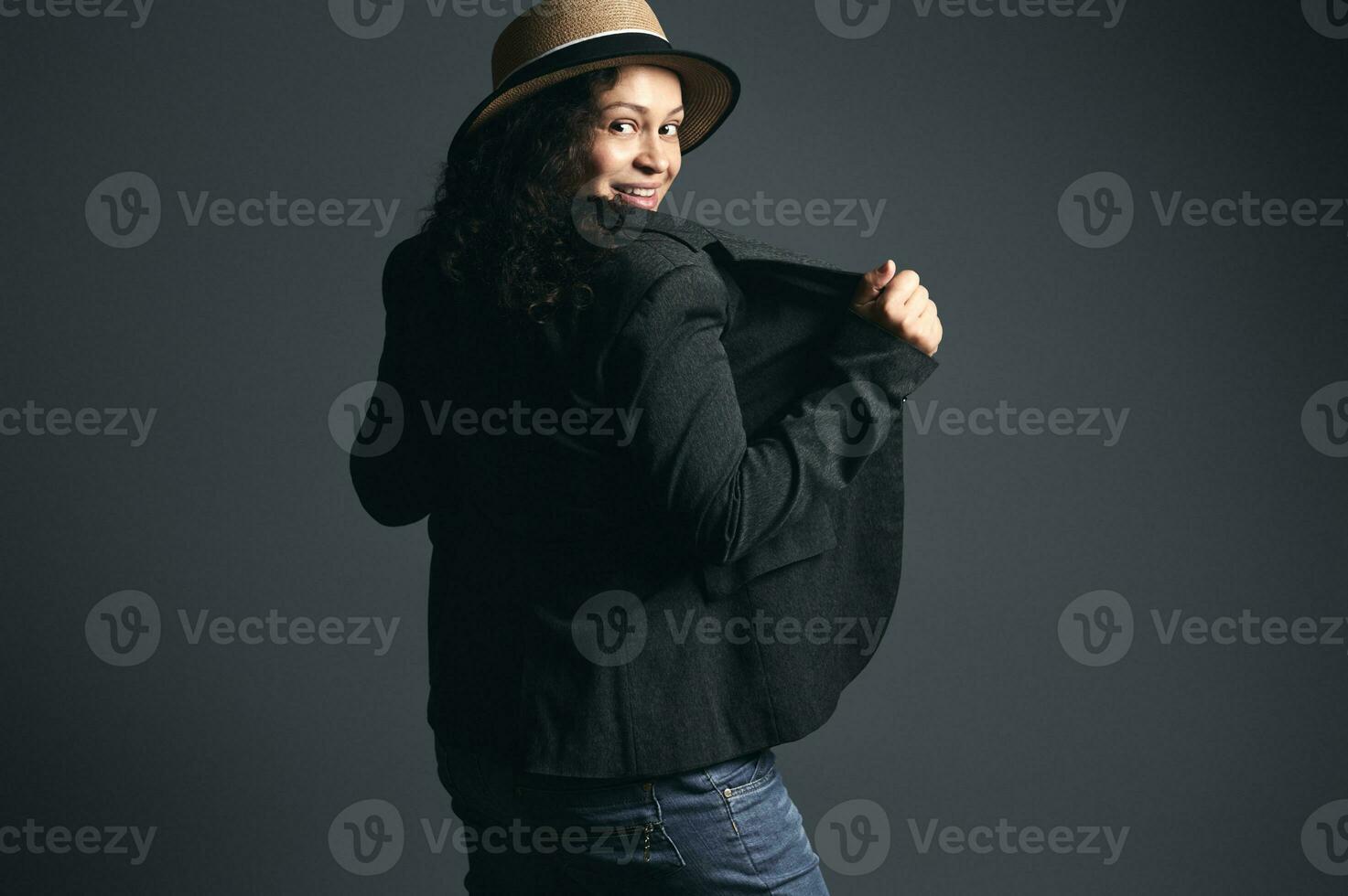 elegante encaracolado latina mulher dentro Palha chapéu e à moda blazer, olhando através dela ombros, posando costas para Câmera foto