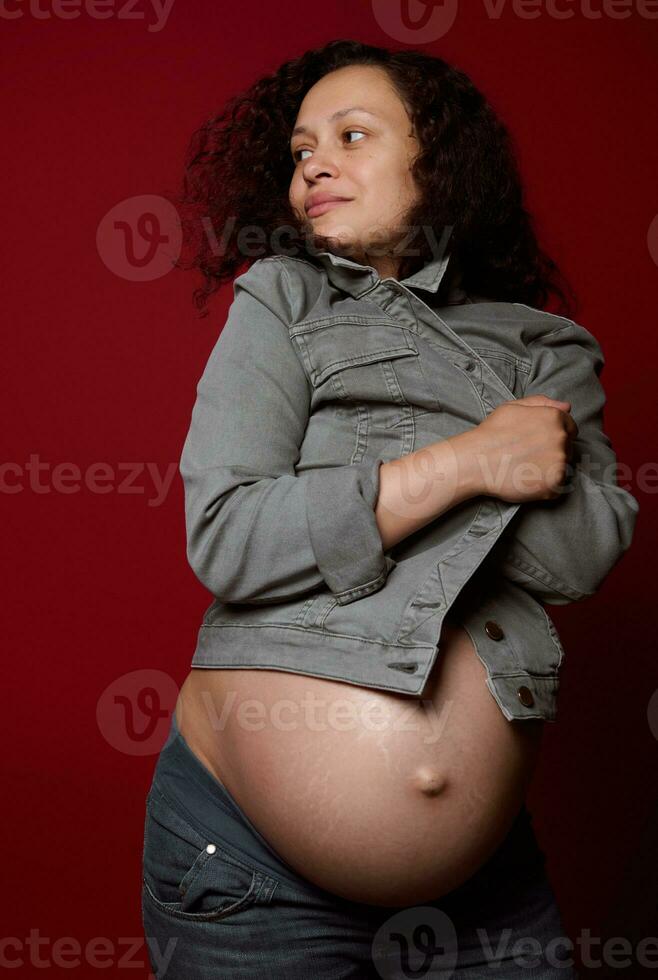 atraente africano americano jovem grávida mulher com nu barriga, vestindo à moda casual jeans blazer, isolado em vermelho foto