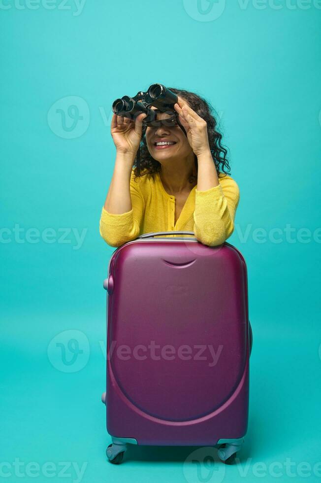 jovem lindo mulher dentro brilhante amarelo roupas inclinado em uma mala de viagem e sorrisos cheio de dentes sorrir olhando através binóculos, isolado em azul fundo com cópia de espaço. explorador, viajante, aventureiro foto