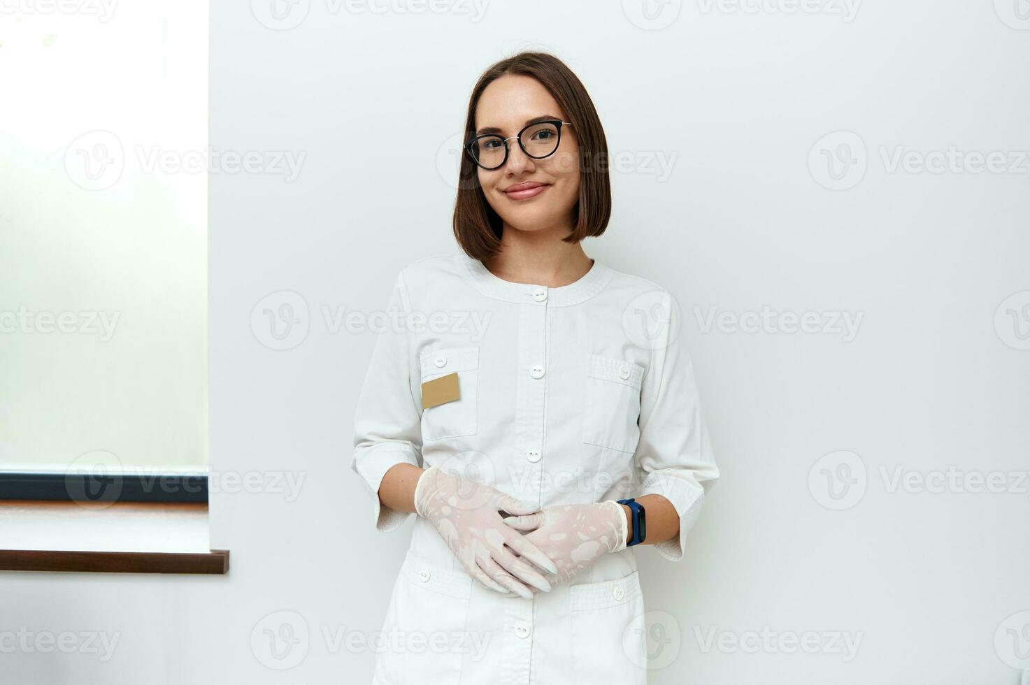 confiante retrato do uma jovem médico dentro branco médico laboratório casaco e protetora luvas olhando às Câmera enquanto posando contra branco parede dentro cosmetologia clínica foto
