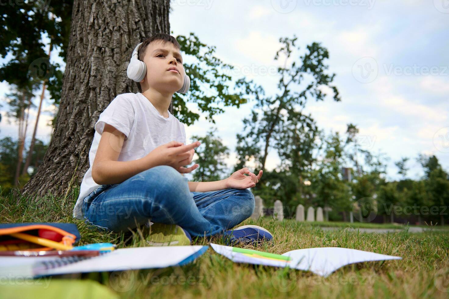 adorável escola criança Garoto sente Exausta e cansado depois de escola e trabalho de casa, senta dentro lótus posição e medita com sem fio fones de ouvido em cabeça. livros de exercícios e escola suprimentos deitado em a Relva foto