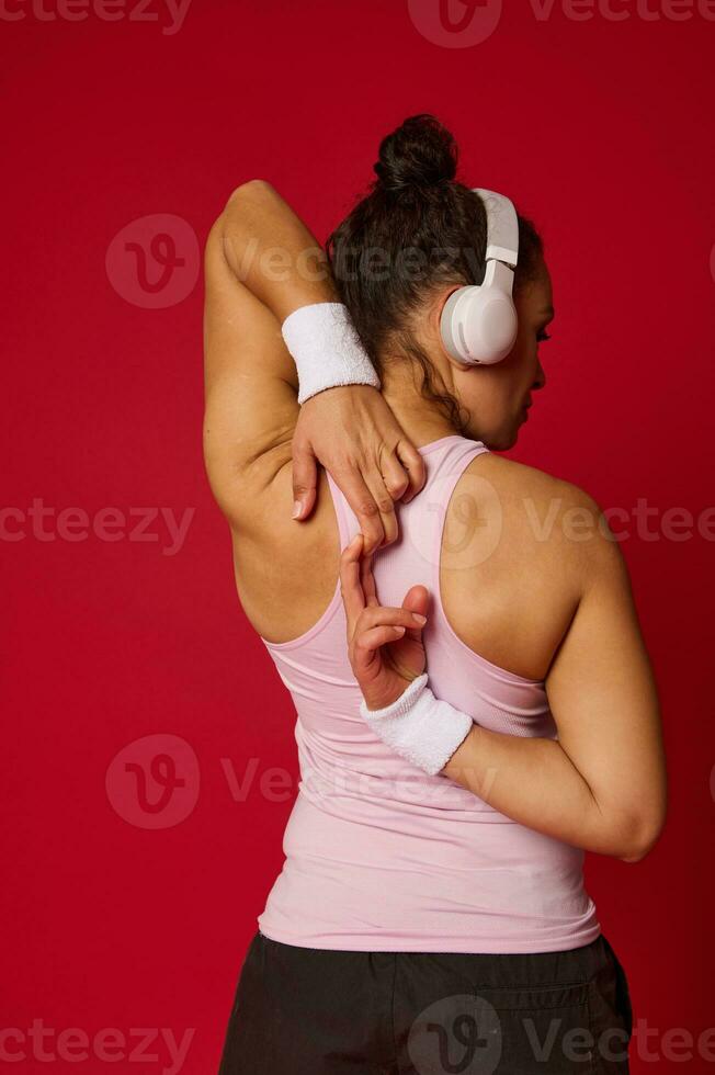 retrato do uma desportista dentro roupa ativa e sem fio fone de ouvido fazendo alongamento exercícios em braços em pé costas para a Câmera contra vermelho fundo foto