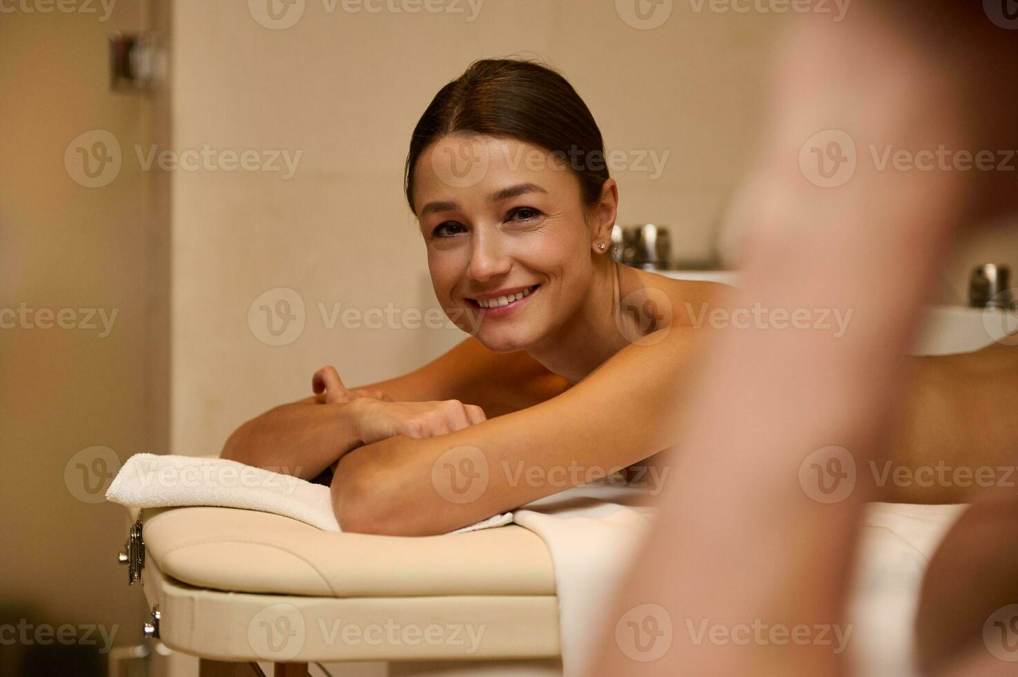 Deleitado meio envelhecido europeu mulher deitado em uma massagem mesa, sorrisos olhando às Câmera, obtendo pronto para receber profissional corpo massagem terapia às bem estar Centro. recém-casados casal às saúde spa foto