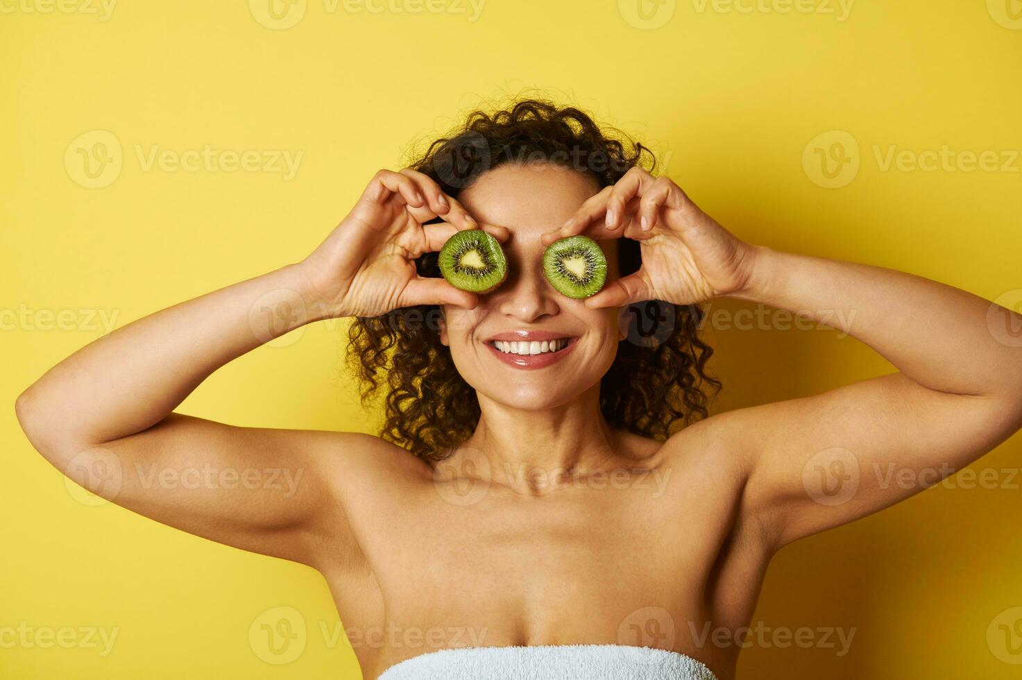 sorridente misturado raça muscular mulher cobertura dela olhos com kiwi metades, sorridente enquanto posando contra amarelo fundo foto