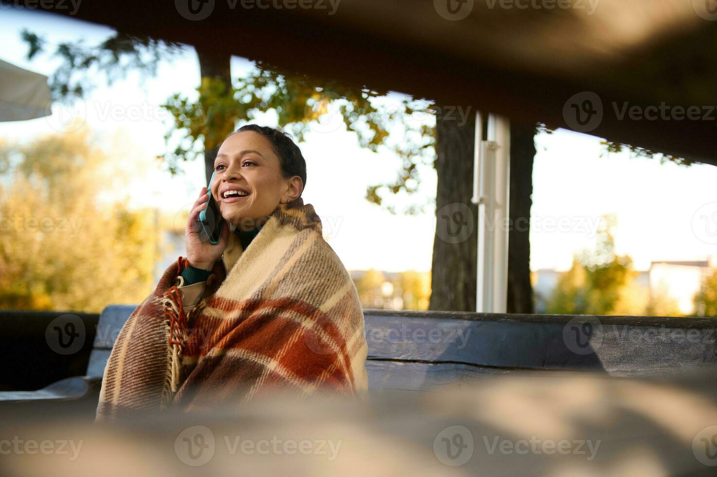 Visão através de madeira Histórico do Banco às sorridente bonita mulher olhando longe enquanto falando em Móvel telefone, sentado em de madeira Banco embrulhado dentro acolhedor xadrez de lã cobertor guardando caloroso em legal outono dia foto