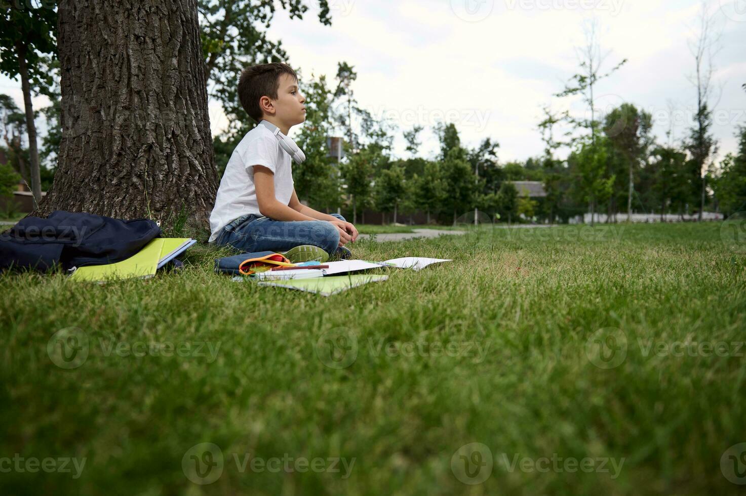 lado Visão para a sereno esperto adorável escola criança Garoto sente Exausta e cansado depois de escola e trabalho de casa, senta dentro lótus posição e medita. livros de exercícios e escola suprimentos deitado em a Relva foto