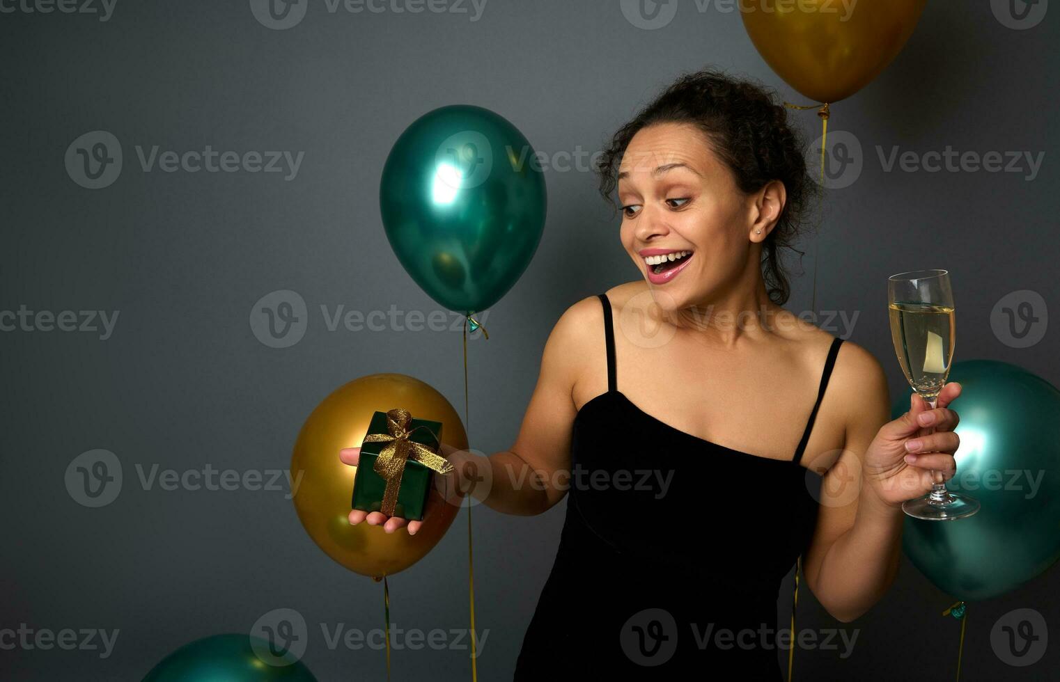 encantador jovem mulher com uma vidro do champanhe dentro dela mãos fofinho alegria às a Natal presente, posando em uma cinzento fundo decorado com balões do ouro e verde metálico cor, cópia de espaço foto