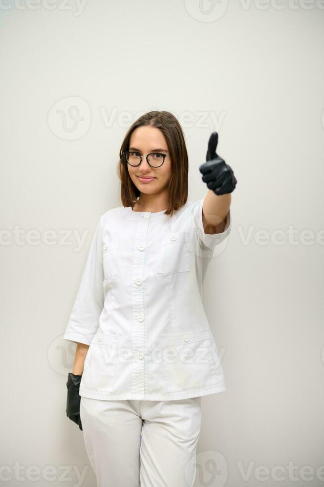 lindo jovem morena mulher, fêmea médico dentro branco uniforme mostrando polegar acima contra branco parede fundo com cópia de espaço foto