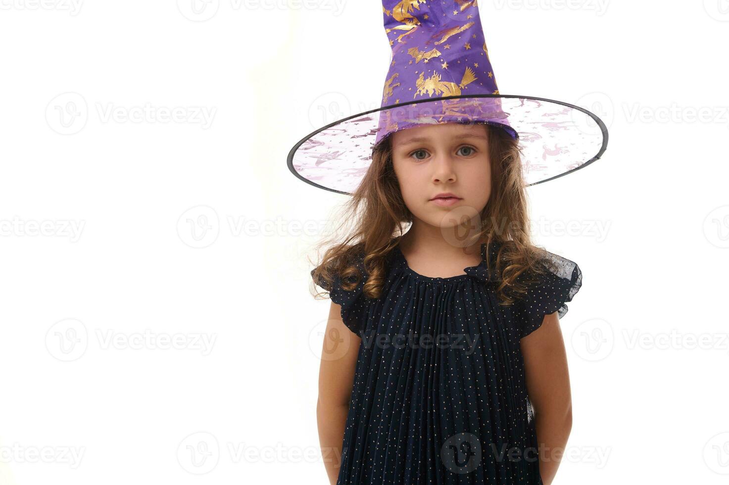 retrato do lindo confiante pequeno menina vestindo uma Mago chapéu e vestido dentro à moda carnaval vestir, olhando às Câmera posando com cruzado braços contra branco fundo, cópia de espaço. dia das Bruxas foto