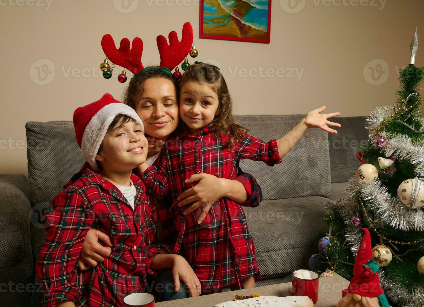 feliz mãe com chifre aro abraços dela adorável crianças vestido dentro vermelho e verde xadrez roupas e santa chapéu em dela filhos cabeça, a comemorar Natal festa às casa dentro família círculo foto