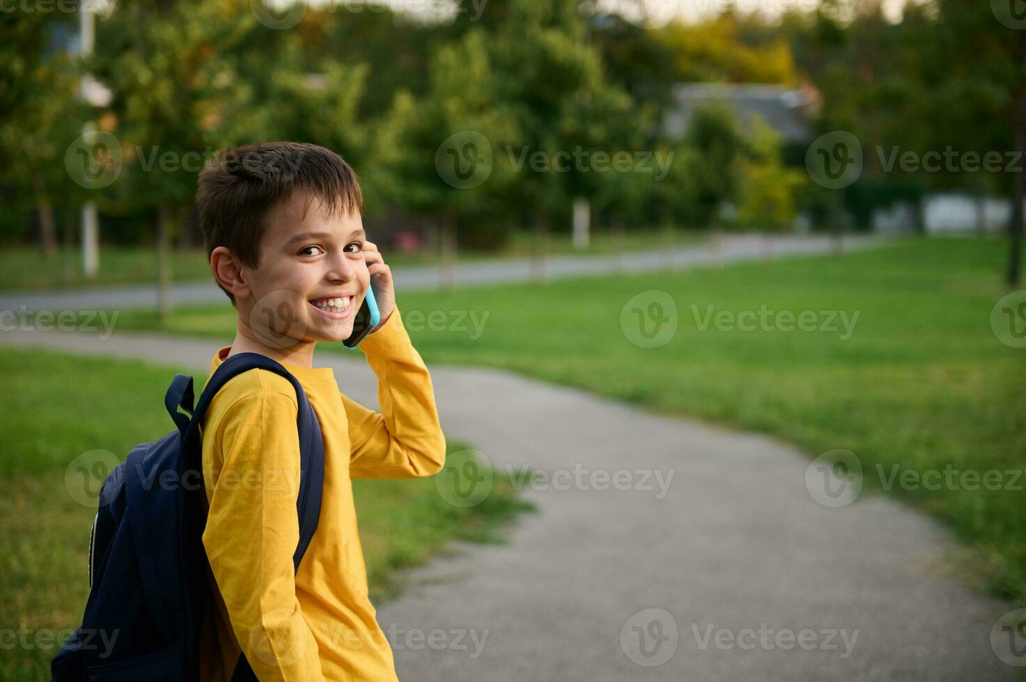 adorável estudante vestindo amarelo suéter com mochila falando em Móvel telefone dentro público parque, indo casa depois de escola, , sorridente com cheio de dentes sorrir para a Câmera foto