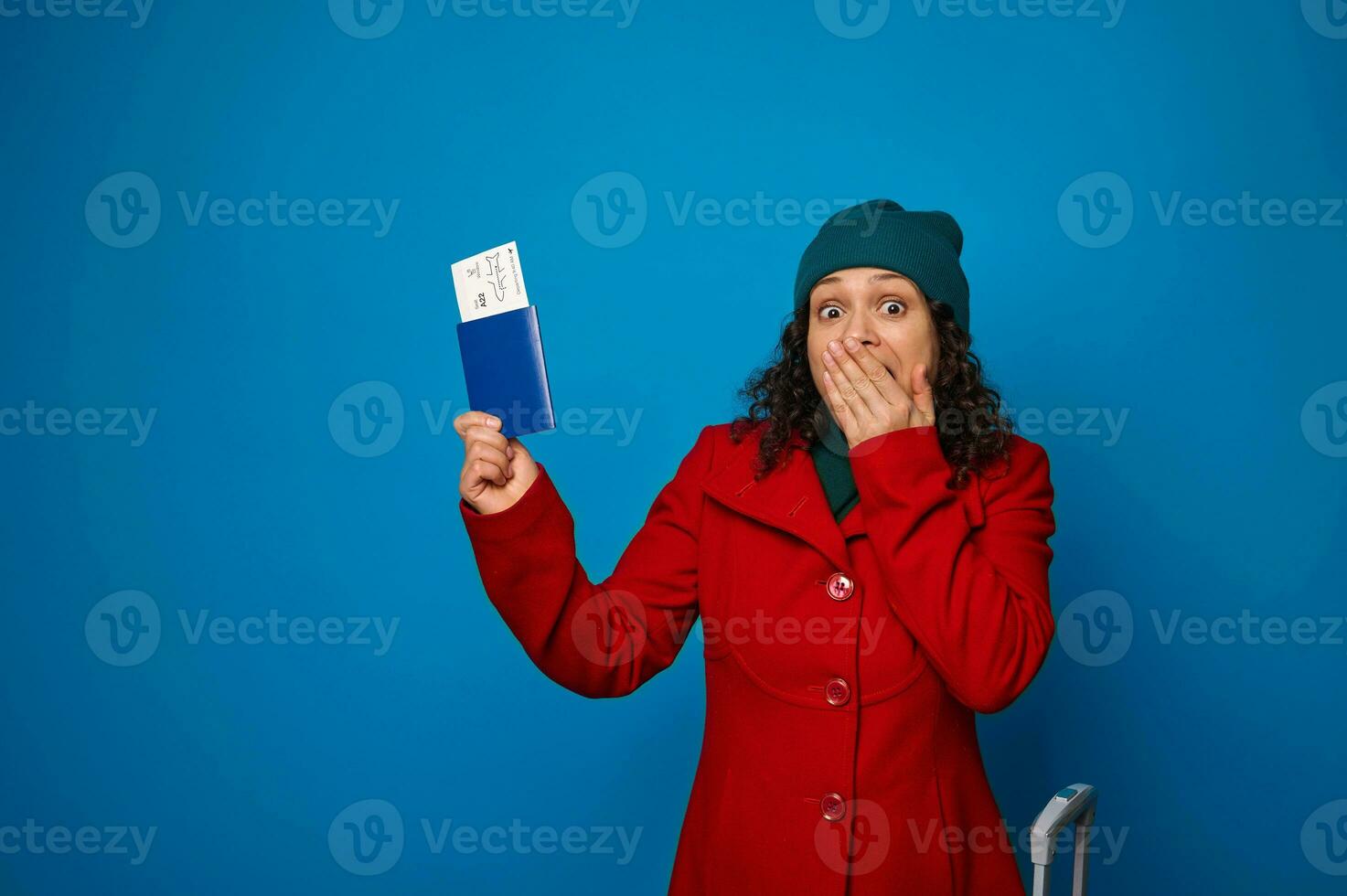 animado atordoado mulher dentro caloroso vermelho casaco e verde chapéu cobre dela boca a partir de surpresa mostrando dela Passaporte com bilhete e embarque passar para a Câmera, isolado sobre azul fundo com cópia de de Anúncios espaço foto