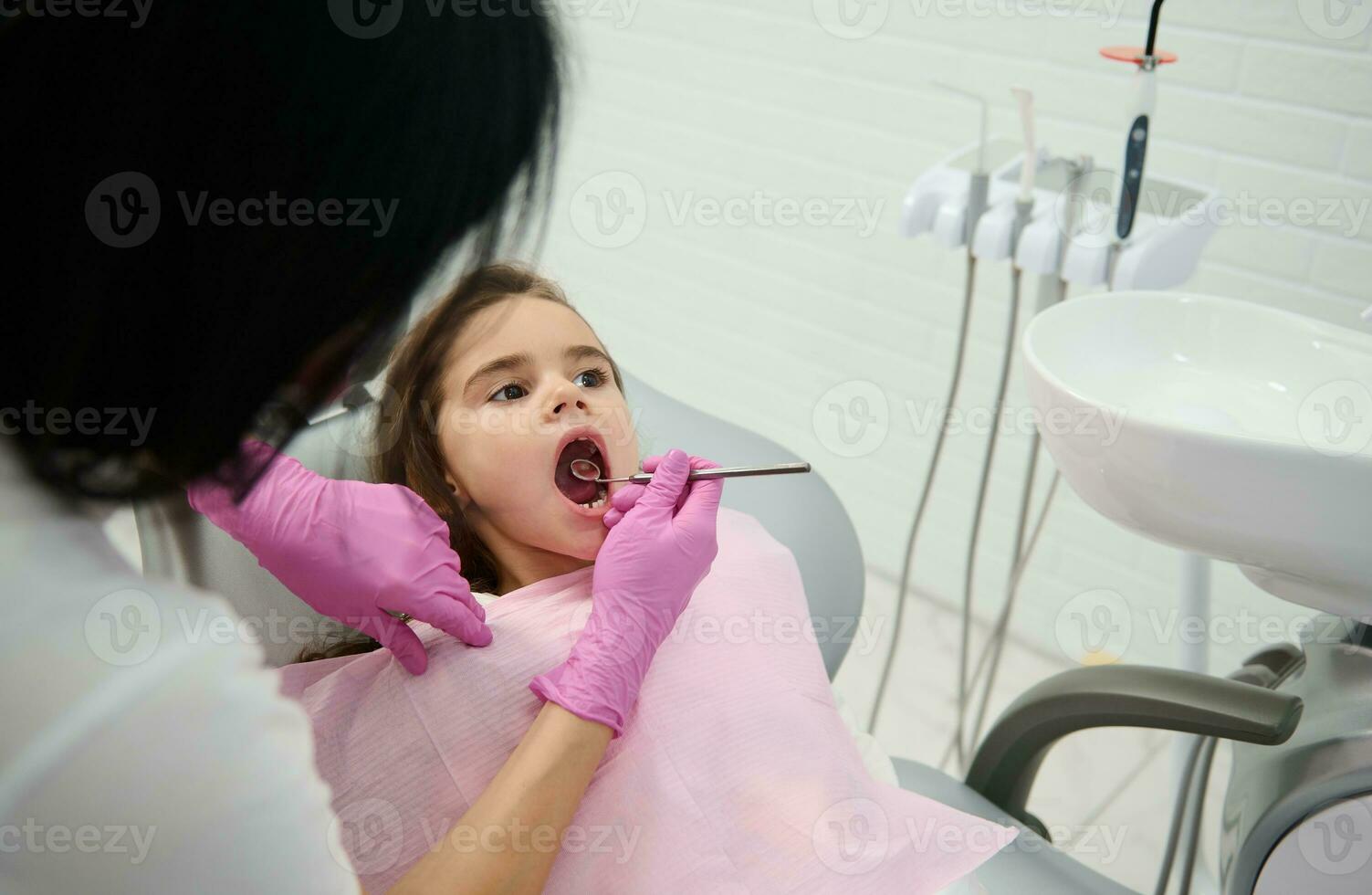 lindo pequeno menina sentado dentro a Dentistas cadeira com dela boca aberto enquanto a pediatra dentista examina dela oral cavidade usando uma inoxidável aço dental espelho dentro uma branco moderno dentista escritório foto