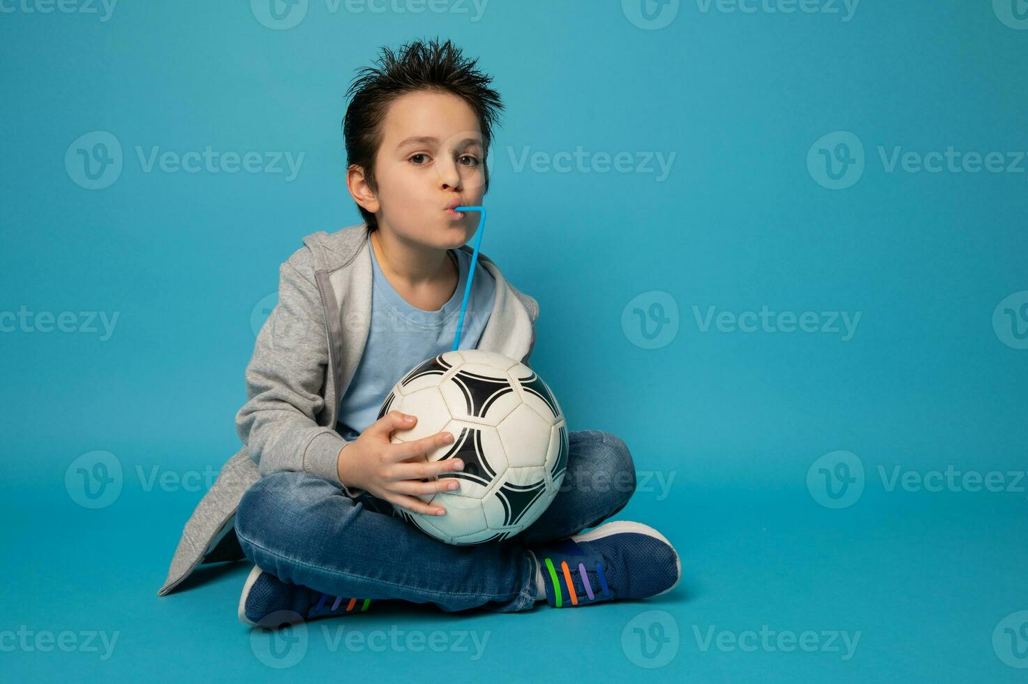 bonito Garoto sentado em azul fundo , segurando uma futebol bola e bebendo a partir de uma Palha foto