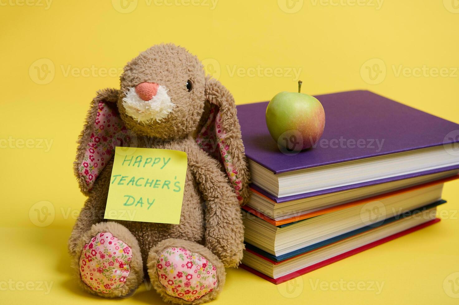 uma pelúcia brinquedo Coelho com letras feliz professores dia em amarelo Nota papel, inclinou-se em uma atiçar do colorida livros, amarelo fundo com espaço para texto foto