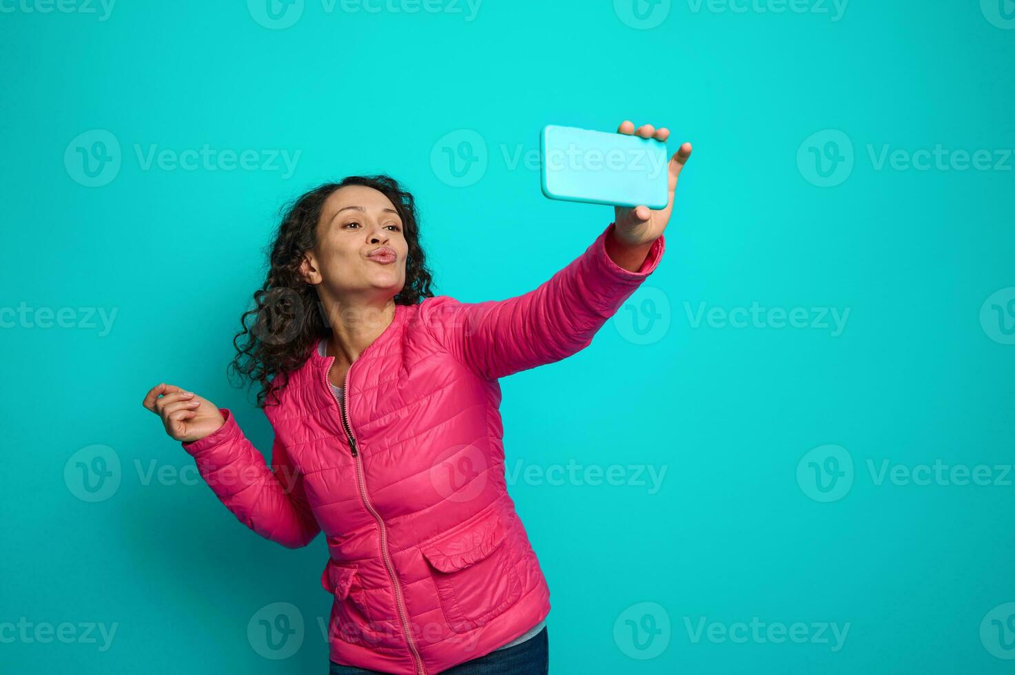 atraente alegre lindo cabelos escuros mulher com encaracolado Penteado, vestindo Rosa jaqueta, envia Beijos olhando às Smartphone Câmera, fazer selfie contra brilhante azul fundo com cópia de de Anúncios espaço foto