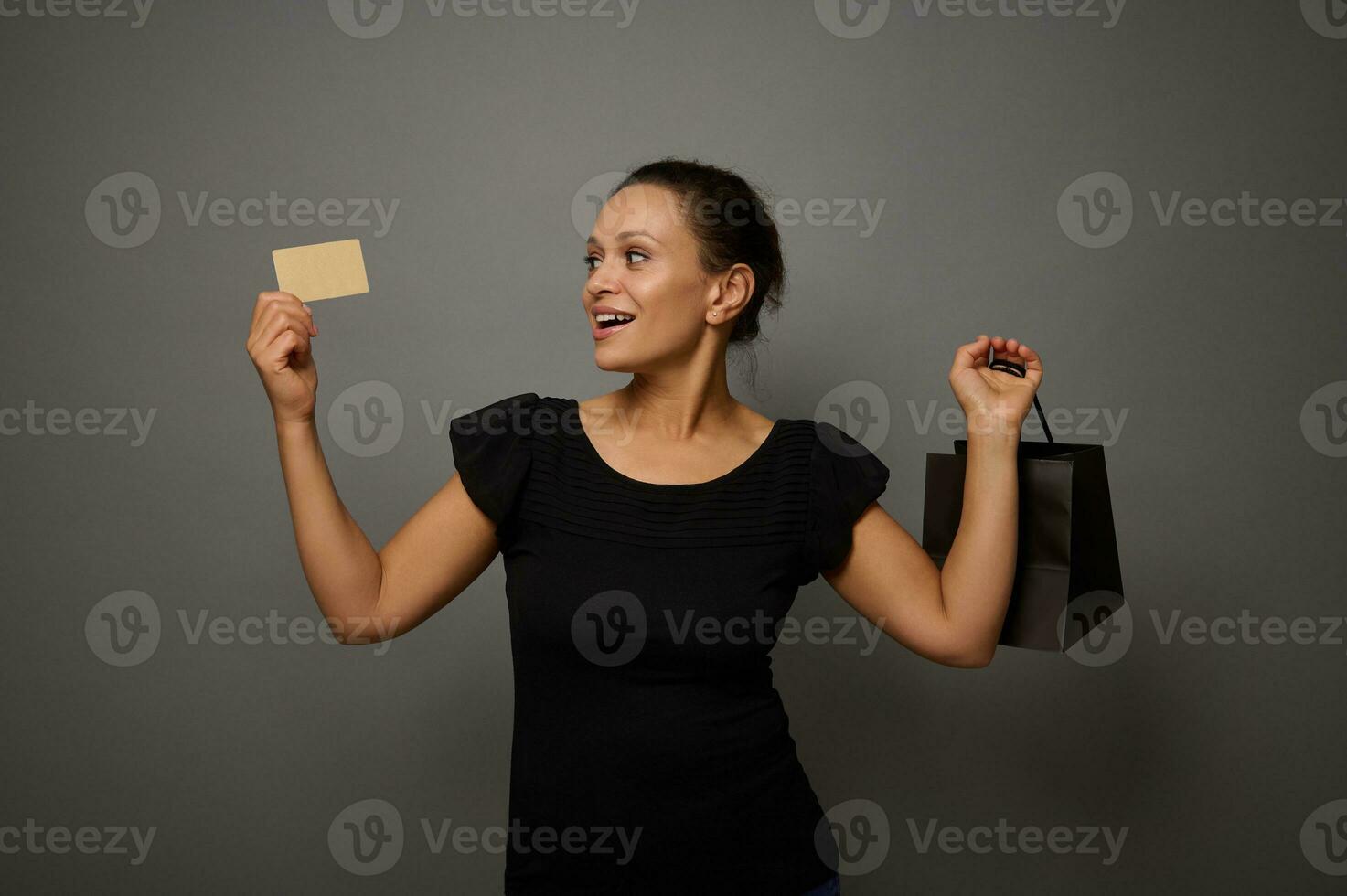 atraente atônito misturado raça lindo mulher poses contra cinzento parede fundo com uma compras pacote e regozija-se olhando às dourado crédito cartão dentro dela mão. Preto Sexta-feira conceito com cópia de espaço foto