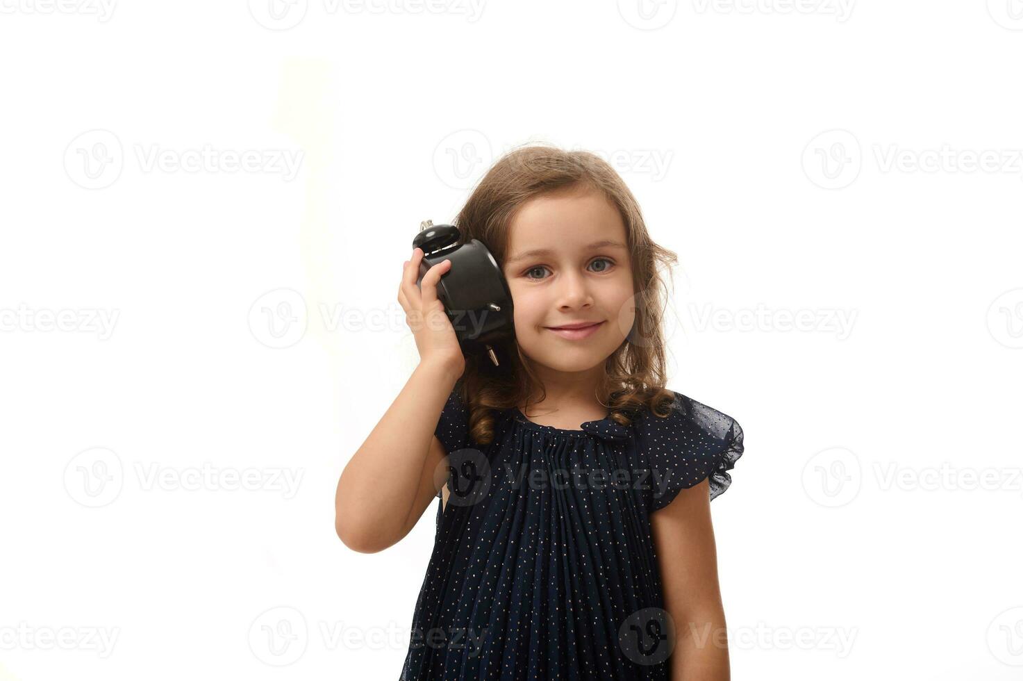 uma lindo 4 anos velho pequeno menina dentro uma Sombrio azul vestir detém a alarme relógio perto dela orelha e escuta atentamente para a som, sorrisos olhando às Câmera, isolado em branco fundo com cópia de espaço foto