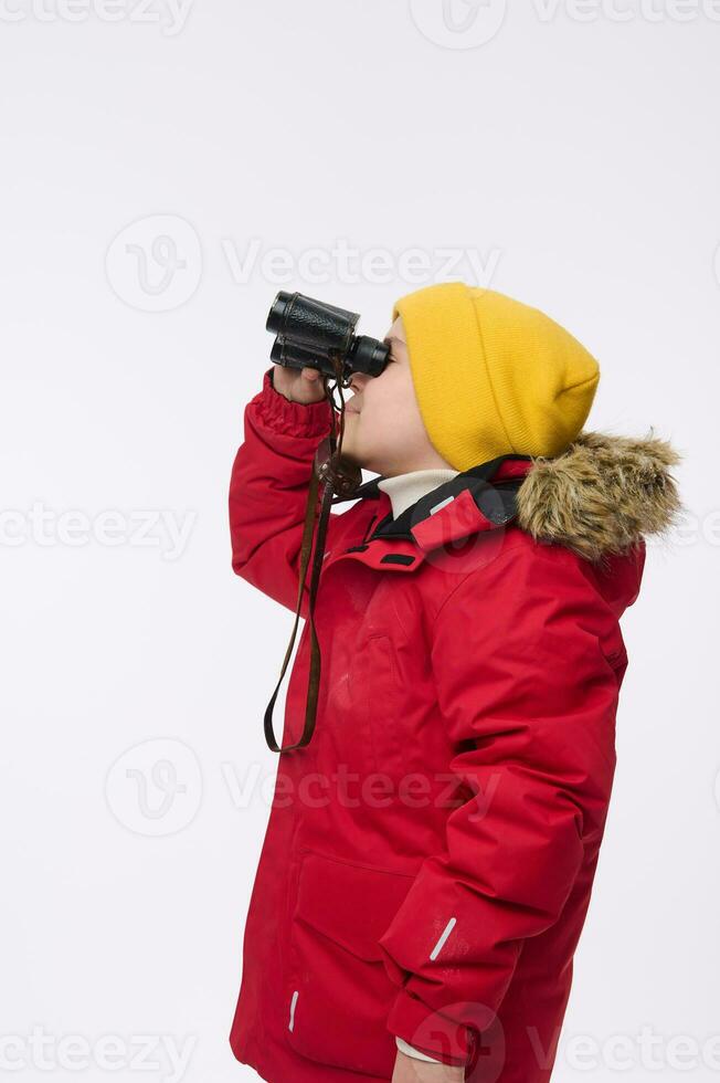 lado retrato do escola Garoto dentro amarelo de lã chapéu e brilhante vermelho baixa jaqueta, olhando através binóculos, isolado em branco fundo, cópia de espaço para anúncio. inverno viagem e aventura conceito foto