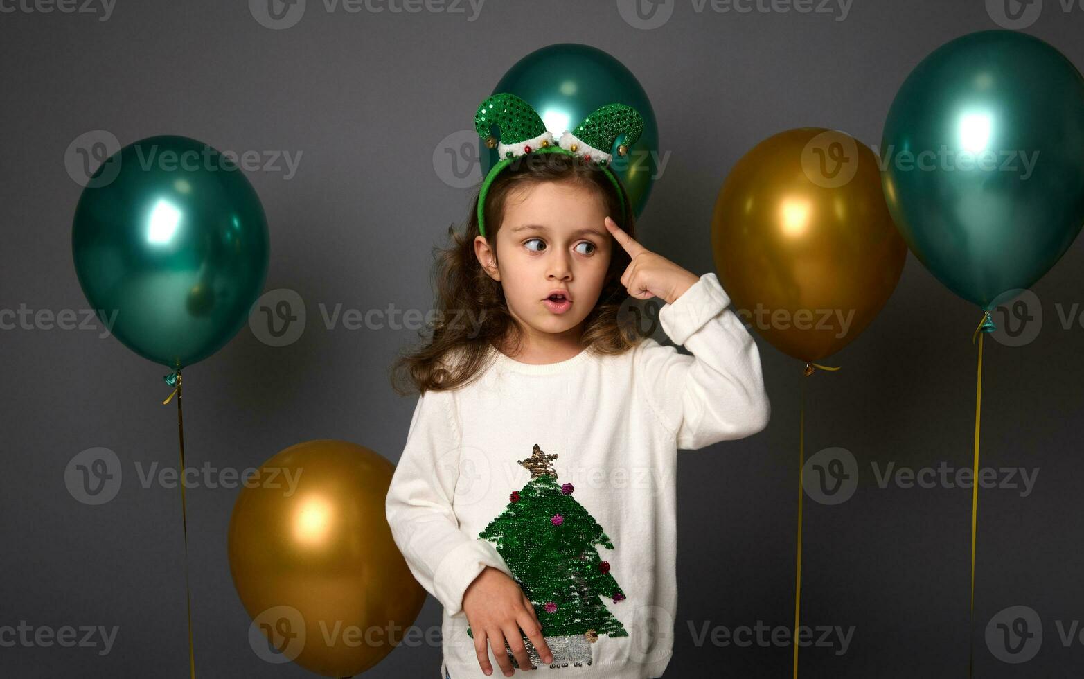 pensativo menina coloca dela dedo em dela têmpora e pensativamente parece abaixo, posando contra cinzento fundo com lindo dourado e brilhante verde metálico ar balões com cópia de espaço para Natal de Anúncios foto