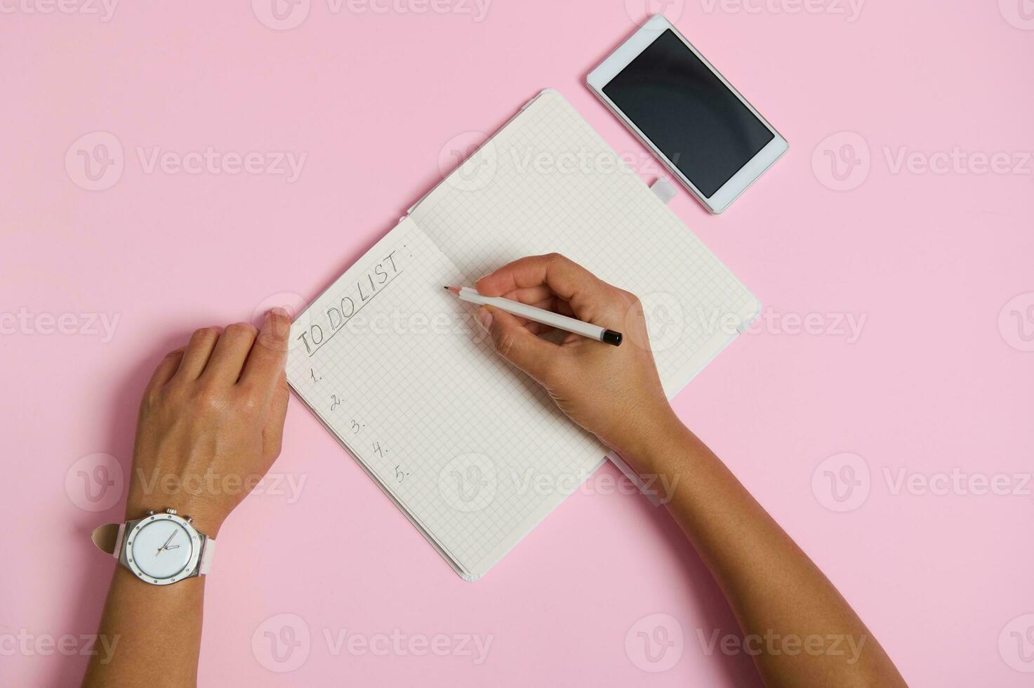 Alto ângulo Visão do fêmea mãos segurando lápis, escrevendo dentro caderno, verificação façam lista. Móvel telefone deitado em Rosa fundo com espaço para texto foto