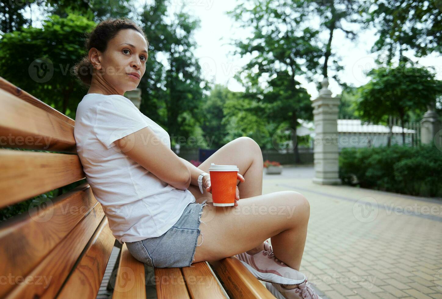 relaxado misturado raça bonita mulher com Leve embora café dentro descartável papel caneca sentado em de madeira Banco dentro a parque foto