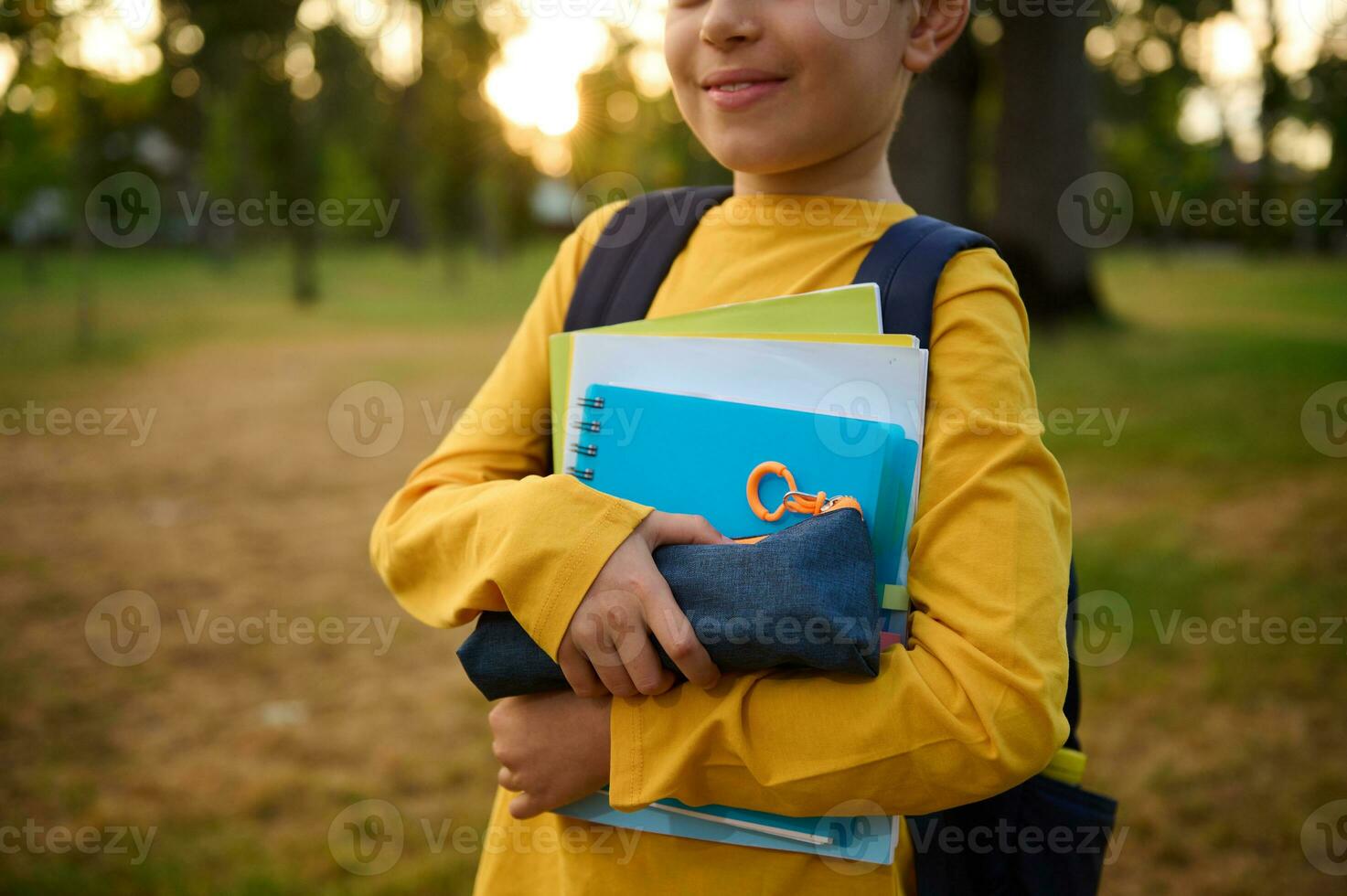 cortada Visão do adorável sorridente escola criança Garoto com escola suprimentos e livros de exercícios dentro a mãos posando para a Câmera dentro a cidade parque às pôr do sol foto