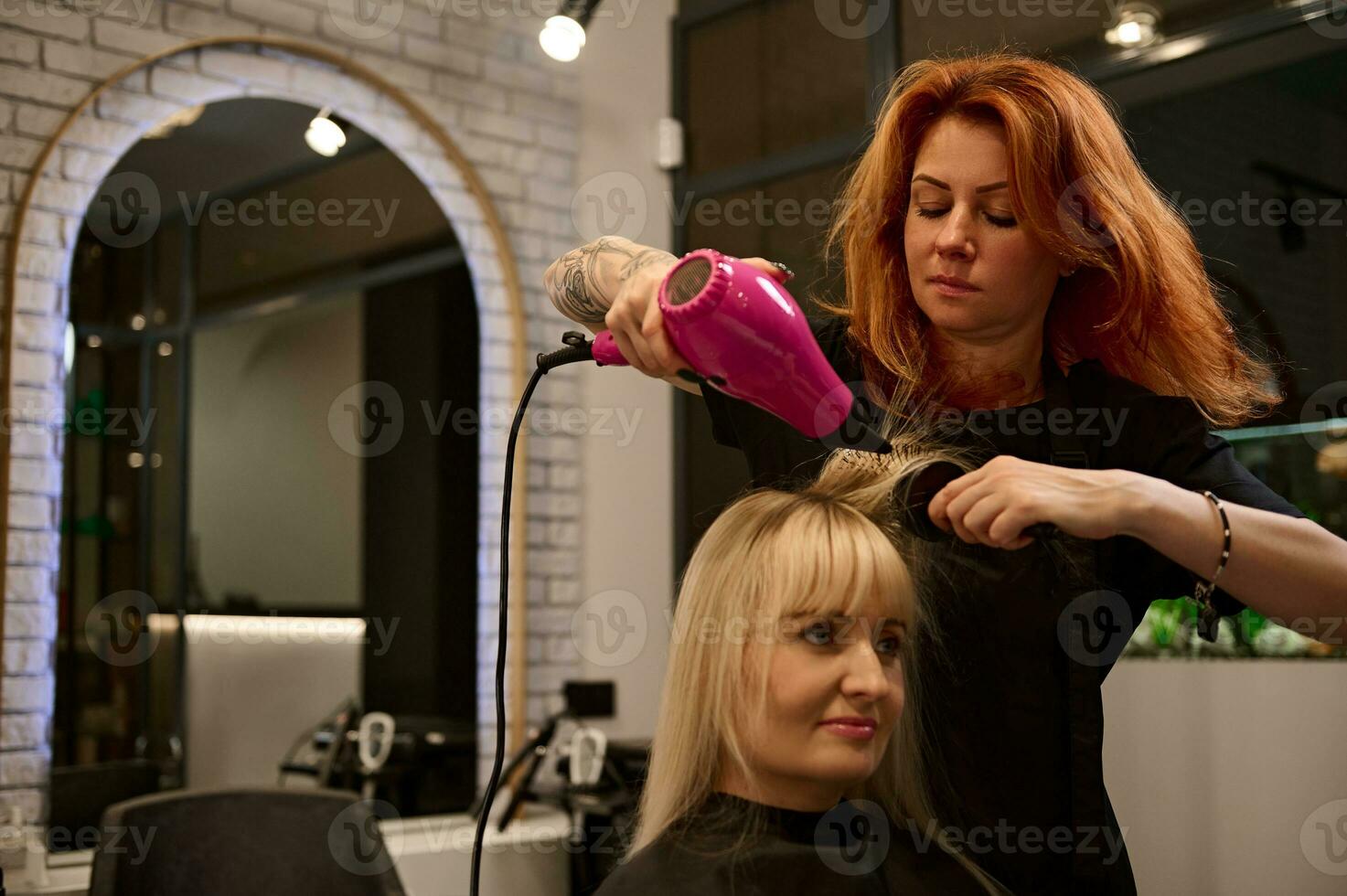 atraente ruiva mulher profissional cabeleireiro com tatuado braços dentro uma rigoroso Preto uniforme pentear, endireitar e secagem a Loiras cabelo do beleza salão cliente usando uma Rosa secador de cabelo foto