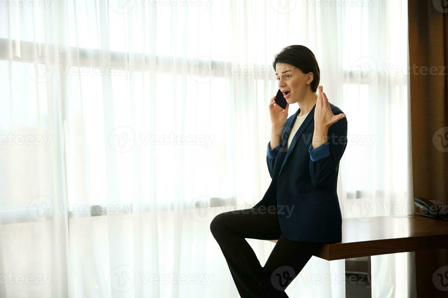 empresária, fêmea funcionário, jovem empreendedor jura, argumenta, gestos com dela mãos enquanto falando em a telefone, sentado às a Beira do uma mesa de janela do uma hotel quarto durante uma o negócio viagem foto