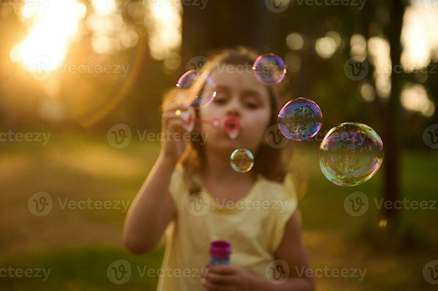 suave foco em Sabonete bolhas em a borrado fundo do uma fofa bebê menina sopro Sabonete bolhas dentro Prado, desfrutando despreocupado infância, lazer em a natureza fundo às pôr do sol foto