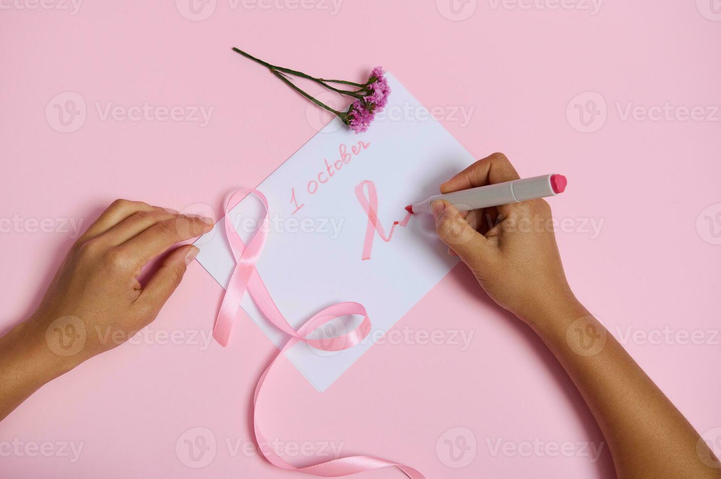 plano deitar do uma mulher mão segurando uma ponta de feltro caneta escreve Outubro 1 e desenha em a papel uma Rosa símbolo do seio Câncer consciência mês, uma Rosa fita com a sem fim fim, deitado em uma Rosa fundo foto