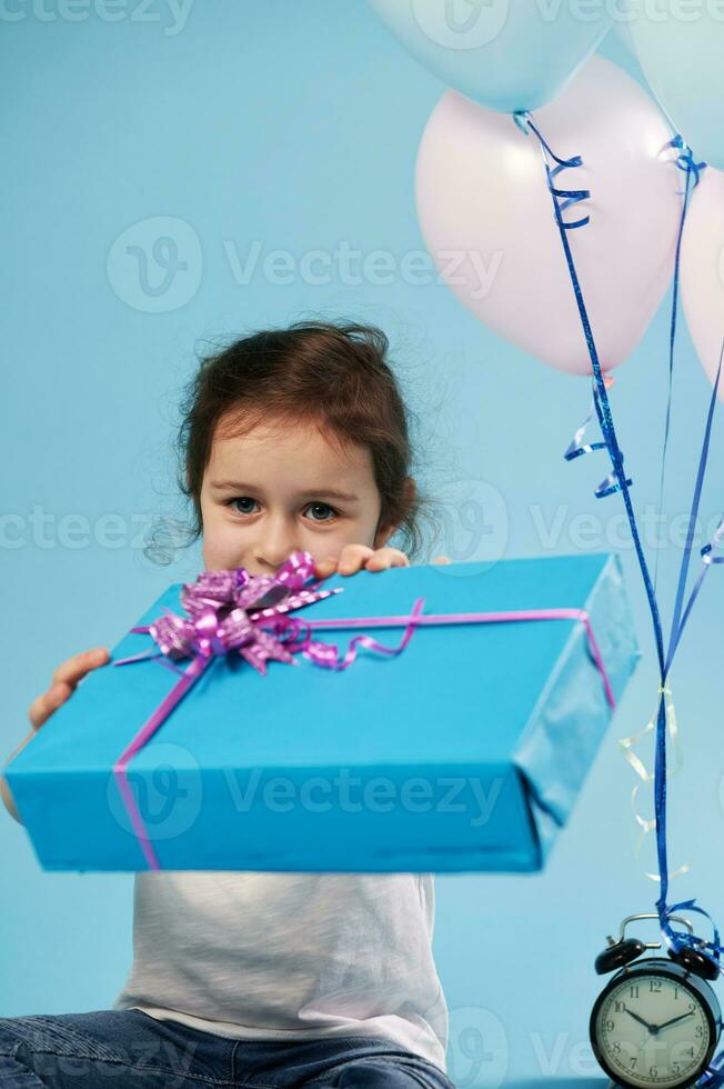 foco em azul presente com uma Rosa arco, que uma fofa pequeno menina detém Fora para a Câmera enquanto sentado em uma azul fundo. celebração conceito foto