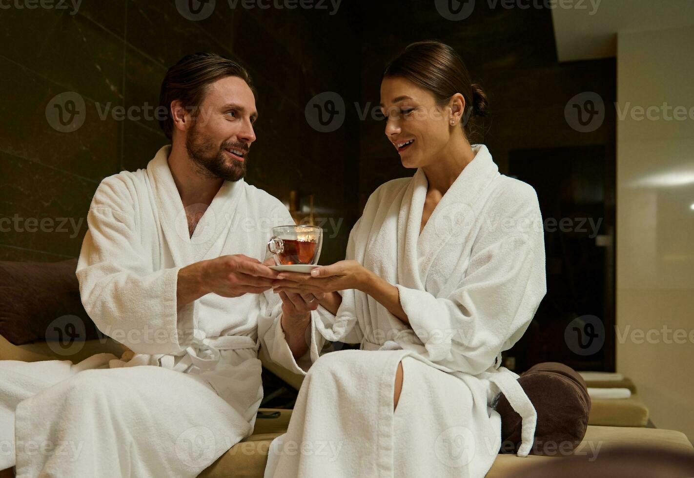 bonito amoroso homem dando uma copo do ervas chá para dela Amado esposa, em repouso juntos dentro salão área de jacuzzi e natação piscina dentro luxo bem estar spa recorrer, desfrutando proximidade em seus lua de mel foto