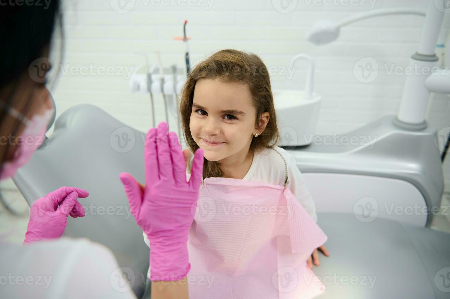 adorável pequeno criança sentado dentro Dentistas cadeira depois de médico checar, sorrisos olhando às Câmera e dá Alto cinco para dela tratando pediatra dentista. lindo menina visitando crianças dentista foto