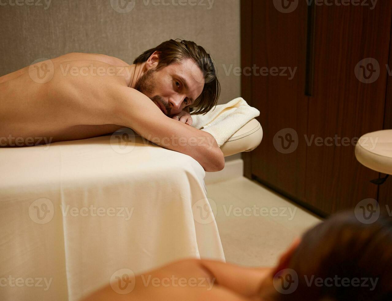uma amoroso casal do meia idade europeu bonito homem, deitado em uma massagem mesa Próximo para dele borrado fêmea parceiro, em repouso juntos dentro uma saúde spa complexo, recebendo ayurvédica corpo Cuidado tratamento foto