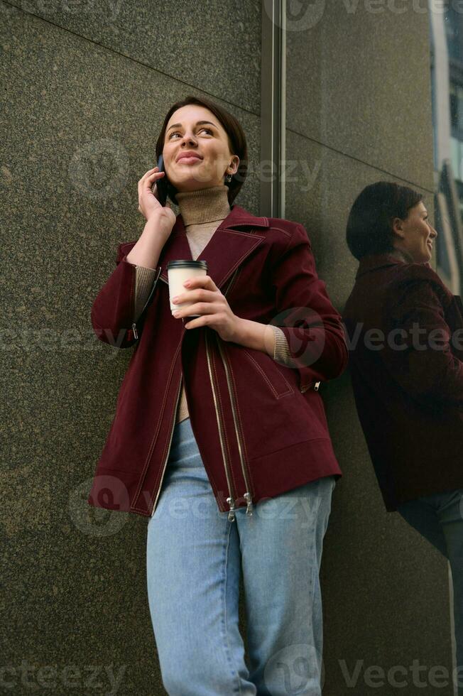 lindo europeu mulher dentro casual vestem fala em Móvel telefone em pé de a mármore parede do uma corporativo construção com uma Leve embora papel copo do café e desfrutando uma café pausa ao ar livre foto