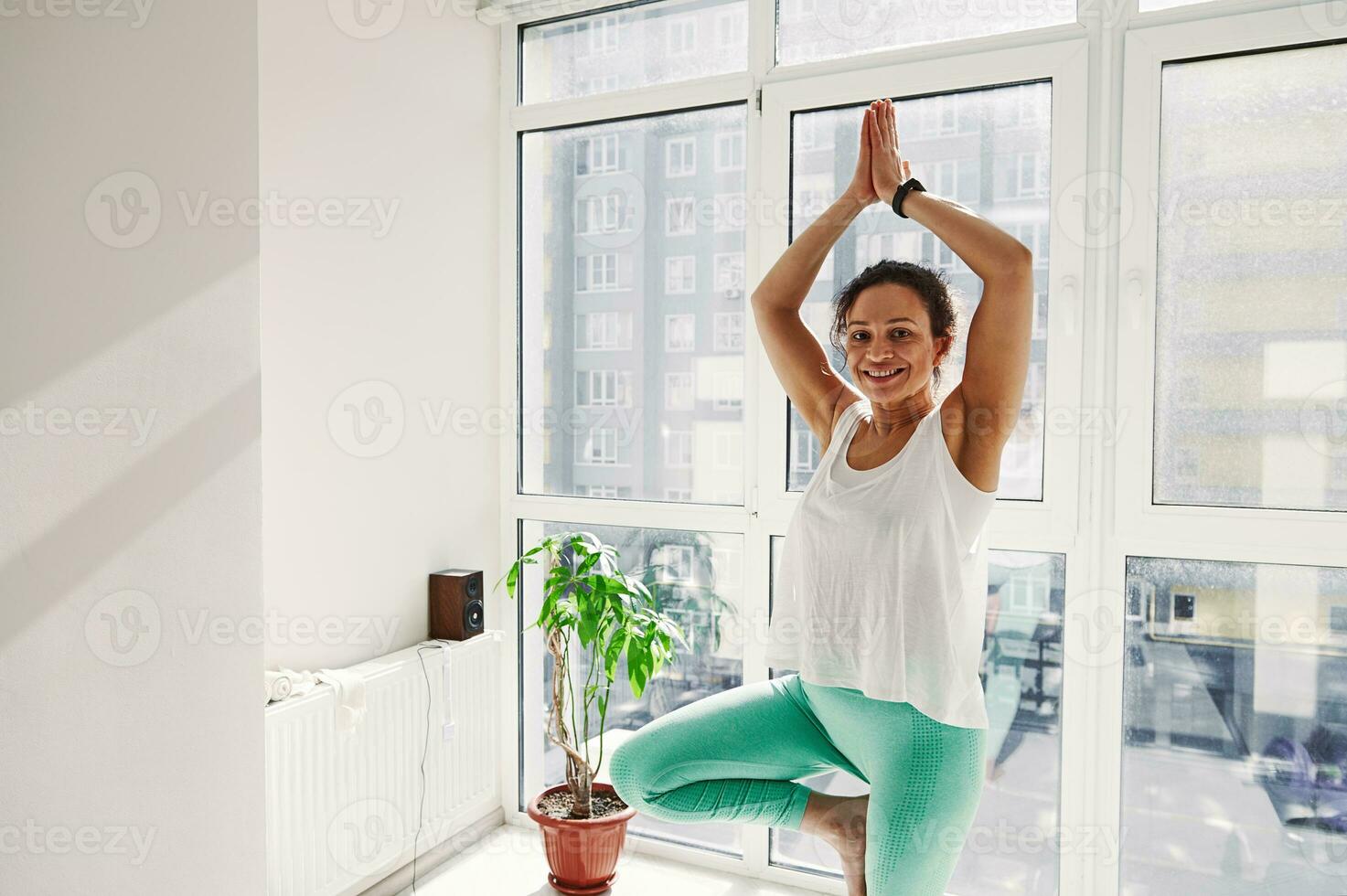jovem Sombrio pele mulher sorrisos às Câmera em pé em árvore posição durante ioga prática foto