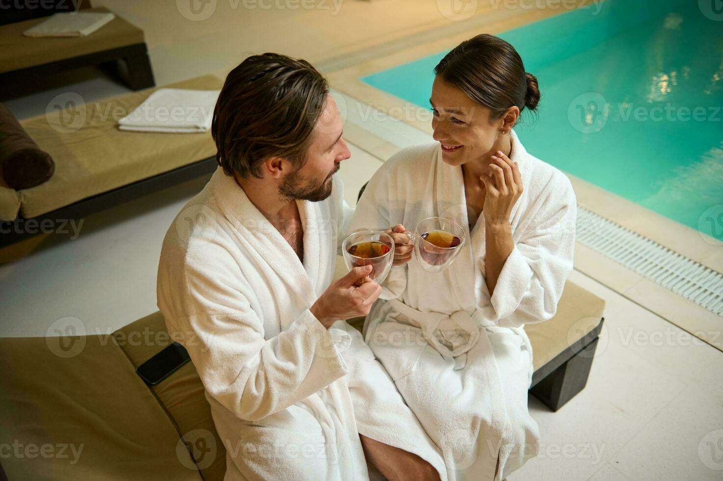 bonito caucasiano homem e linda mulher, vestindo branco Terry roupões de banho, arrepiante, em repouso dentro salão área de piscina dentro uma luxuoso bem estar spa recorrer, fofinho falando sobre copo do saudável ervas chá foto