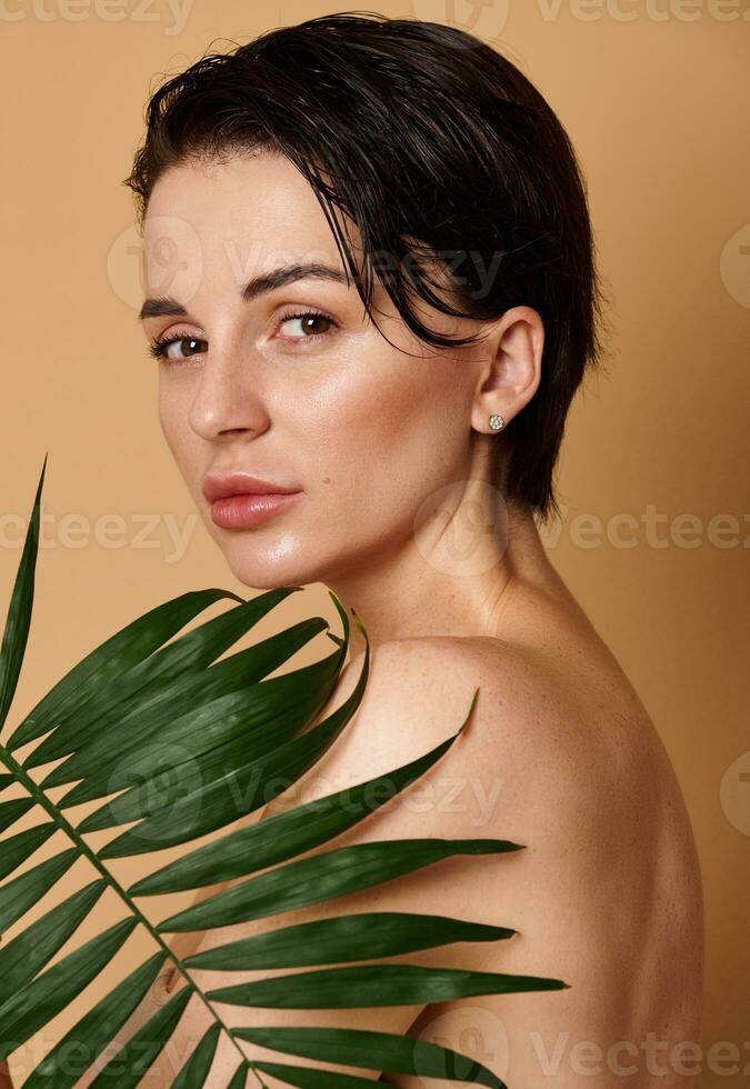 fechar-se retrato do a atraente sensual natural beleza mulher cobertura dela costas com exótico Palma folha, com confiança olhando às Câmera. pele, corpo Cuidado conceito em bege fundo com cópia de de Anúncios espaço foto