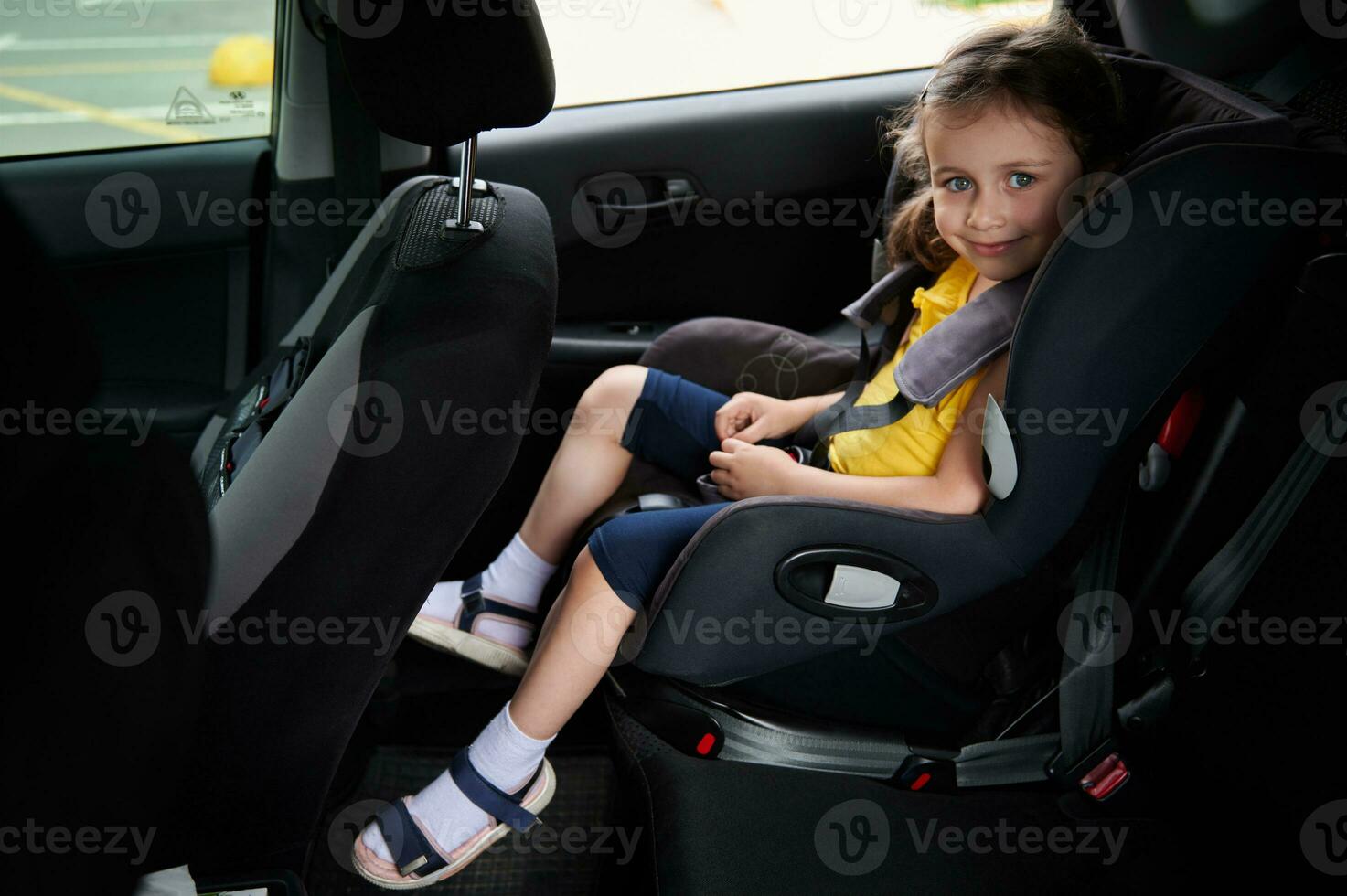 bebê menina com cinto dentro uma segurança criança carro assento, sorrisos enquanto olhando às a Câmera. usando uma criança carro assento para viagem em segurança com crianças dentro uma carro foto